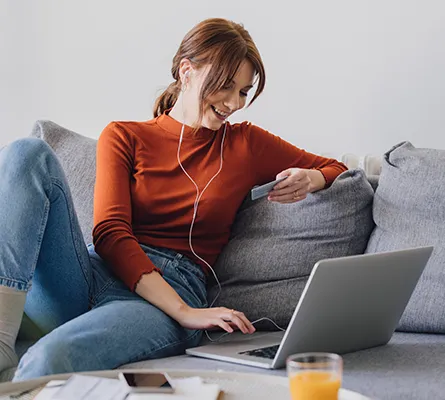 A woman with headphones looking at her credit card in front of a laptop.